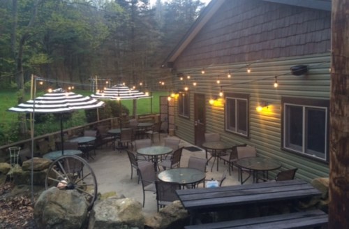 Outdoor patio with tables, chairs, and striped umbrellas, illuminated by string lights, surrounded by greenery.