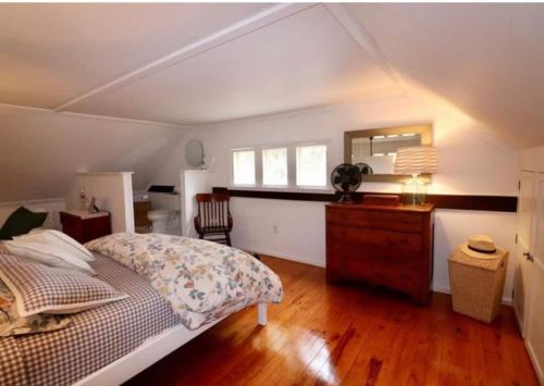 Cozy attic bedroom with a bed, dresser, chair, and natural light from windows. Warm wooden floors and simple decor.
