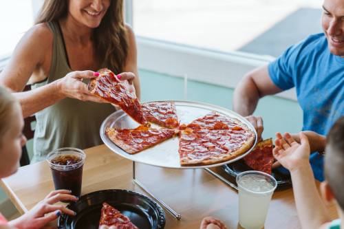 A family enjoys pizza at a table, with one person serving slices from a large pizza on a tray. Drinks are nearby.