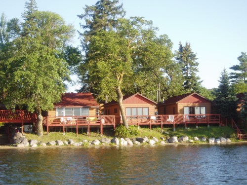 Three cozy cabins with red decks by a serene lake, surrounded by lush green trees and a clear blue sky.