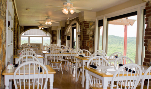 Bright dining area with wooden tables and white chairs, featuring large windows and scenic views of greenery outside.