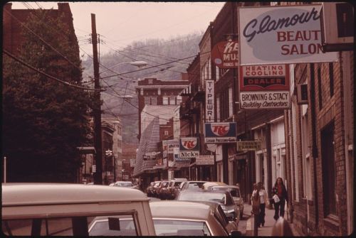 A vintage street scene featuring shops and signs, with cars parked along the road and mountains in the background.