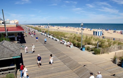 A sunny beach boardwalk with people walking, lined with benches and beach umbrellas, overlooking the ocean.