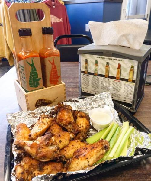 A tray of crispy chicken wings with celery sticks and dipping sauce, alongside two bottles of hot sauce and a menu holder.