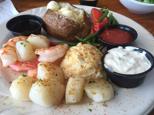 A plate featuring shrimp, scallops, crab cake, baked potato, green beans, and dipping sauces.