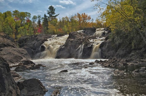 A scenic waterfall cascades over rocky cliffs, surrounded by colorful autumn foliage and a serene river.