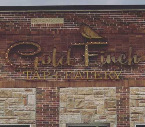 Sign for "Gold Finch Tap Eatery" on a brick wall, featuring gold lettering and a decorative finch illustration.