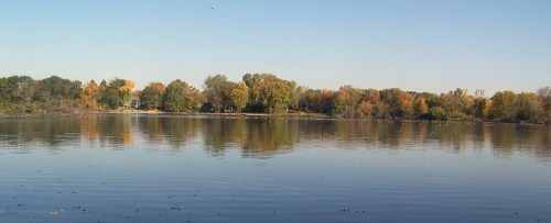 A serene lake surrounded by trees with autumn foliage, reflecting the clear blue sky and calm water.
