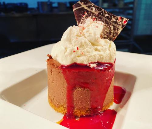A rich chocolate dessert topped with whipped cream, berry sauce, and a chocolate shard, served on a white plate.