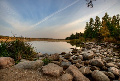 A serene lakeside view with smooth stones, lush greenery, and a clear sky reflecting on the water's surface.