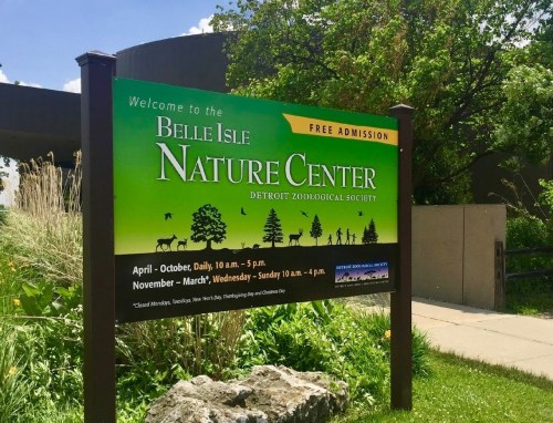 Sign for Belle Isle Nature Center, featuring hours and free admission details, surrounded by greenery.