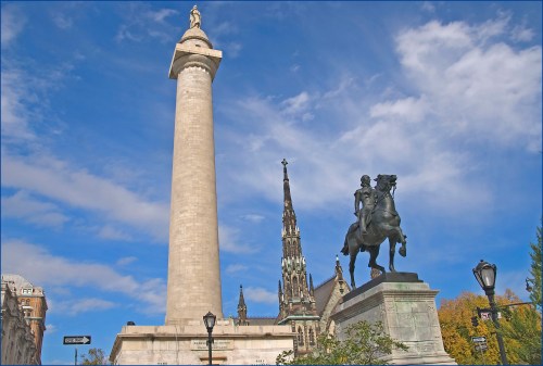 A tall stone monument with a statue on top, alongside a horse statue and a church spire under a blue sky.
