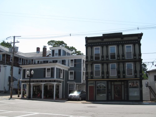 Two historic buildings on a street corner, one with a shopfront and the other a multi-story structure. Clear blue sky.