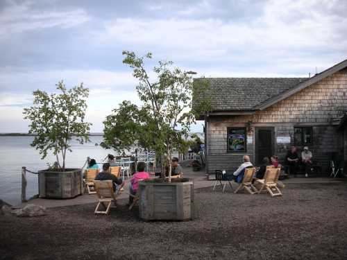 A lakeside scene with people sitting in chairs near a wooden building, surrounded by trees and water.