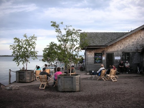 A lakeside scene with people sitting in chairs near a wooden building, surrounded by trees and water.