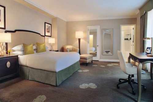 A spacious hotel room featuring a king bed, desk, armchair, and large mirror, with soft lighting and neutral decor.