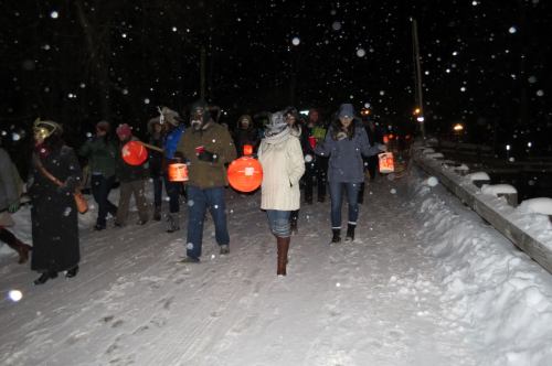 A group of people walking in the snow at night, holding orange lanterns, with snowflakes falling around them.