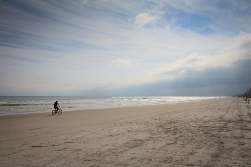 A person riding a bicycle along a sandy beach under a cloudy sky with gentle waves in the background.