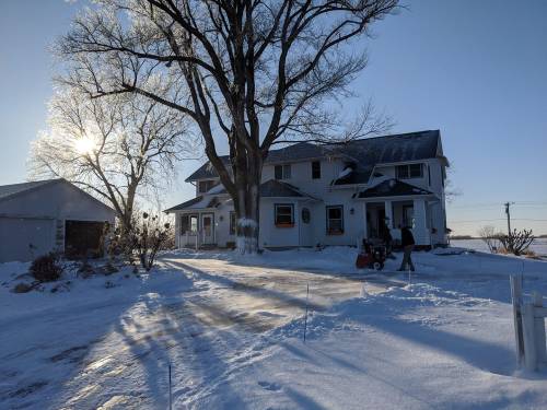 A snowy landscape featuring a large house, a person using a snowblower, and a bright sun in the background.