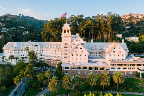 Aerial view of a large, historic hotel surrounded by palm trees and hills, with an American flag flying atop the tower.