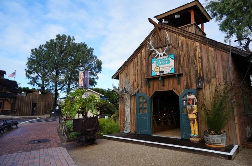 A rustic wooden barn with a sign, surrounded by greenery and a pathway, under a blue sky with scattered clouds.