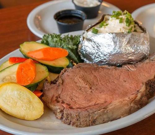 A plate featuring a slice of prime rib, a baked potato with toppings, and a side of mixed vegetables.
