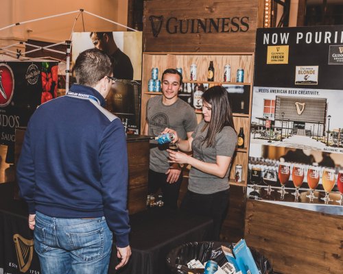 A man interacts with staff at a Guinness booth, showcasing beer cans and promotional materials at an event.