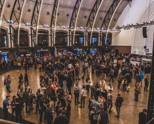 A large crowd gathers in a spacious venue with high ceilings, enjoying an event filled with booths and activities.