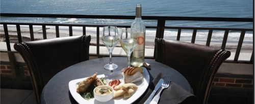 A seaside dining setup with a plate of food, two glasses of wine, and a bottle, overlooking the ocean.