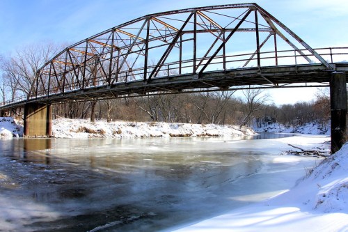 A rusty, old bridge spans a frozen river, surrounded by snow-covered trees under a clear blue sky.