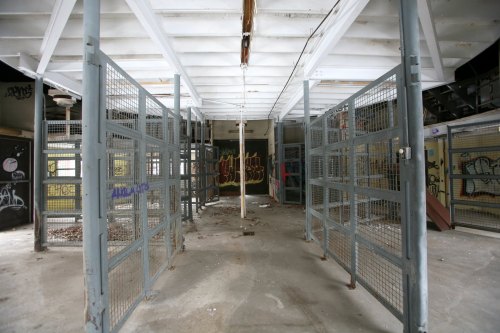 Abandoned interior of a building with metal cages and graffiti on the walls, featuring a high ceiling and concrete floor.