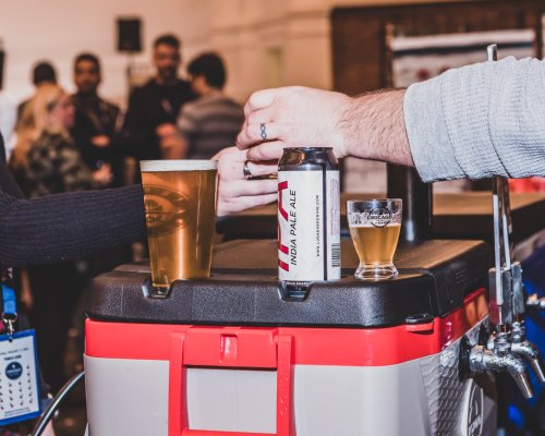 A hand pours a drink from a can into a glass, with a pint and a shot glass on a cooler in a lively setting.