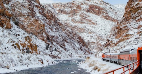 A train travels alongside a snowy river, surrounded by steep, snow-covered mountains.
