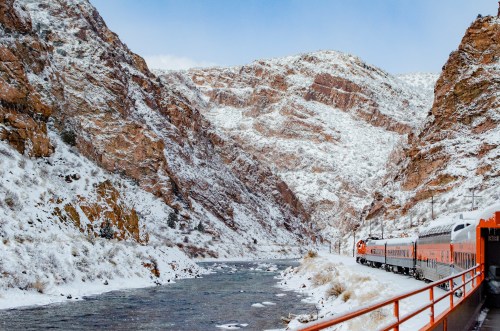 A train travels along a snowy riverbank, surrounded by towering, snow-covered mountains.