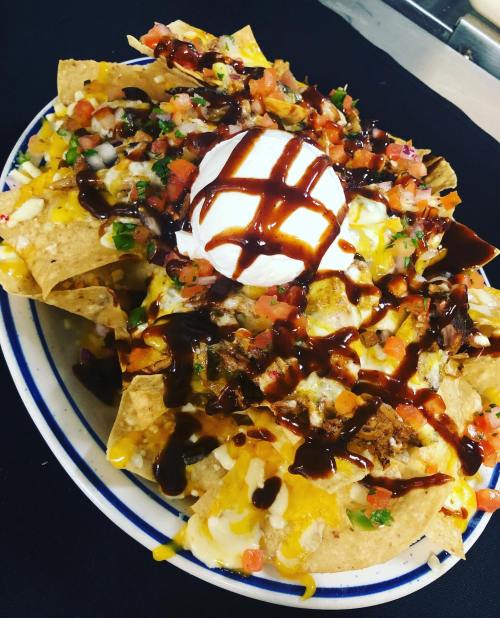 A plate of loaded nachos topped with cheese, pico de gallo, sour cream, and drizzled with barbecue sauce.