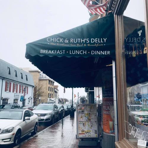 A storefront with an awning reading "Chick & Ruth's Delly," featuring flags and a rainy street scene.