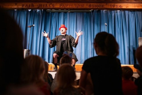 A performer in a top hat and cape entertains an audience from the stage, with children watching in the foreground.