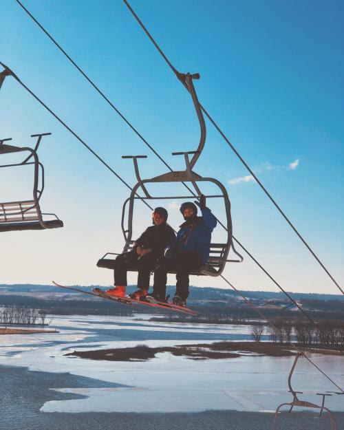 Two people on a ski lift against a clear blue sky, overlooking a snowy landscape and frozen water below.