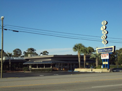 A diner named "Dino's House of Pancakes" with a retro sign and palm trees, set against a clear blue sky.