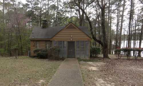 A small wooden cabin surrounded by trees, with a path leading to the entrance and a lake visible in the background.