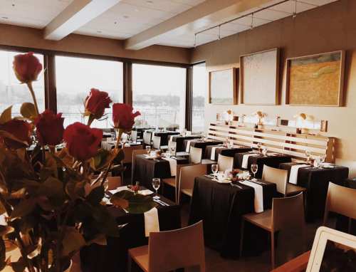 Elegant restaurant interior with tables set for dining, featuring roses in the foreground and large windows with a view.