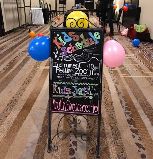 A chalkboard sign displaying a kids' schedule with events like an instrument petting zoo and a youth showcase. Colorful balloons are nearby.