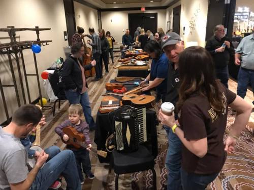 A lively gathering with people playing instruments and socializing at a music event, featuring tables of various musical instruments.
