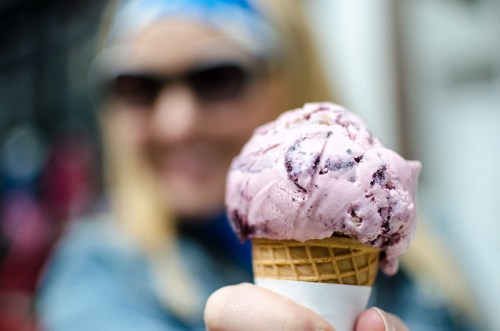 A close-up of a hand holding a cone of pink ice cream, with a blurred person smiling in the background.