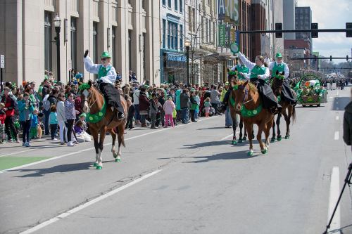 A parade scene with riders in green attire on horseback, celebrating with a crowd in festive attire along the street.