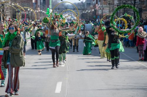 A lively parade scene with participants in green costumes, hula hoops, and festive decorations celebrating a holiday.