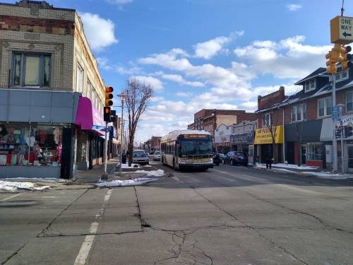 A city street scene with a bus, shops, and traffic lights under a partly cloudy sky. Snow is visible on the ground.