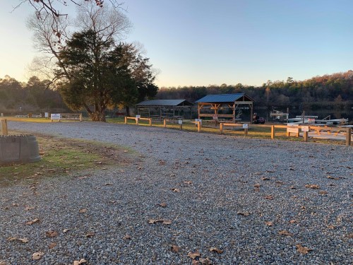 Gravel path leading to a lakeside area with wooden structures and trees in the background during sunset.