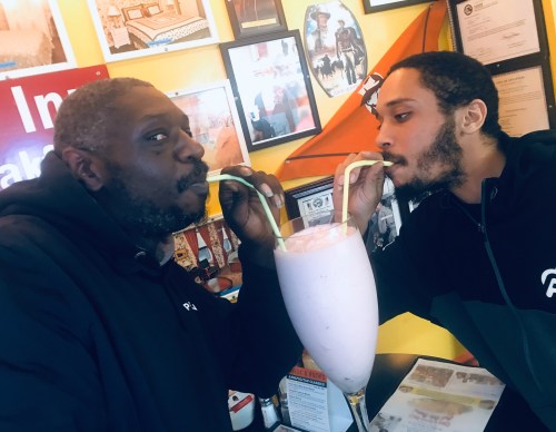 Two men share a large milkshake, sipping from straws, in a colorful diner filled with framed photos.
