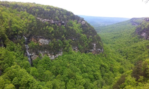 Lush green valley with steep cliffs and a waterfall cascading down, surrounded by dense forest under a cloudy sky.